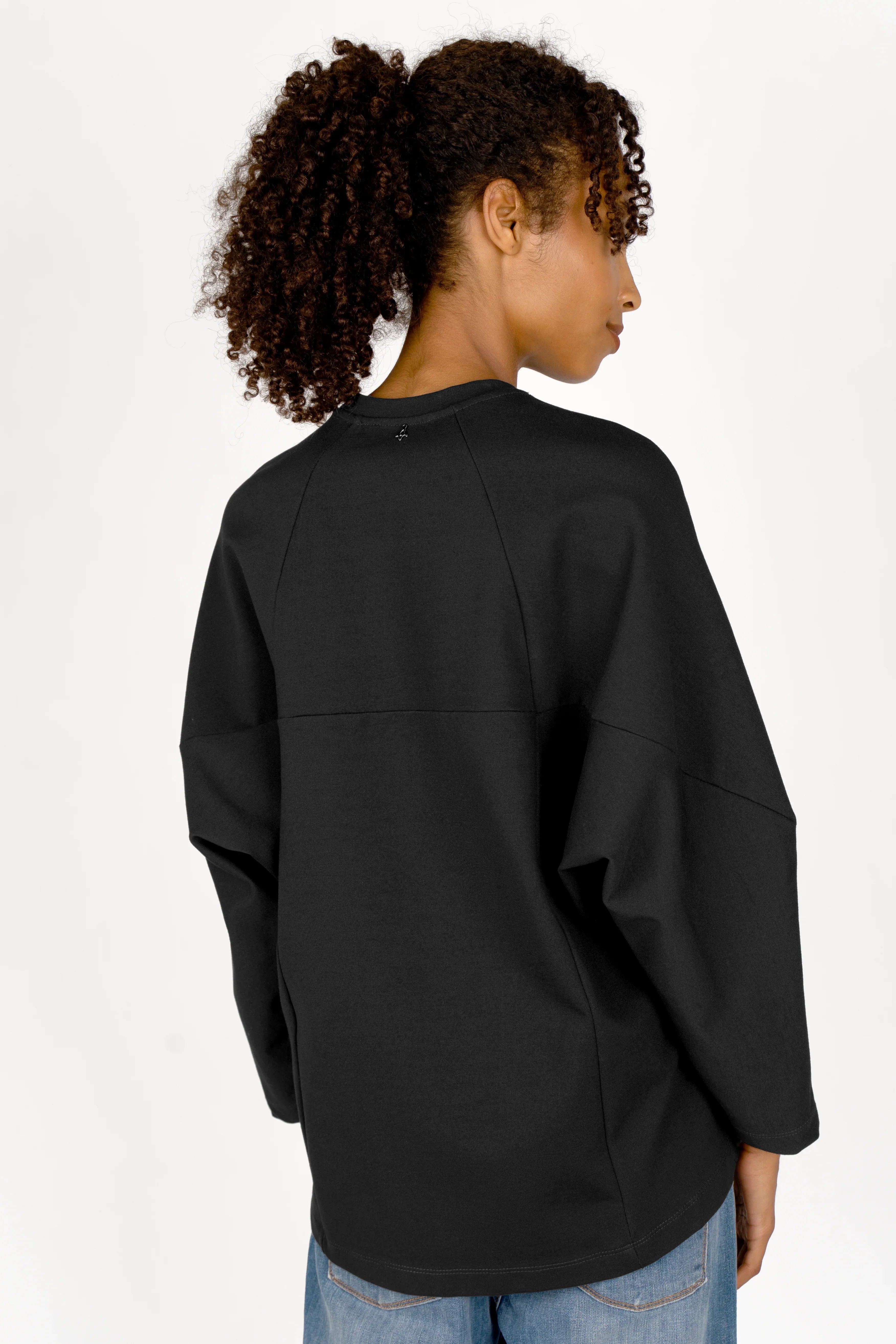 Frau trägt schwarzes TOOVE Shirt aus mittelschwerem Interlock-Jersey mit überschnittenen Schultern und verlängerter Rückenpartie, Rückansicht