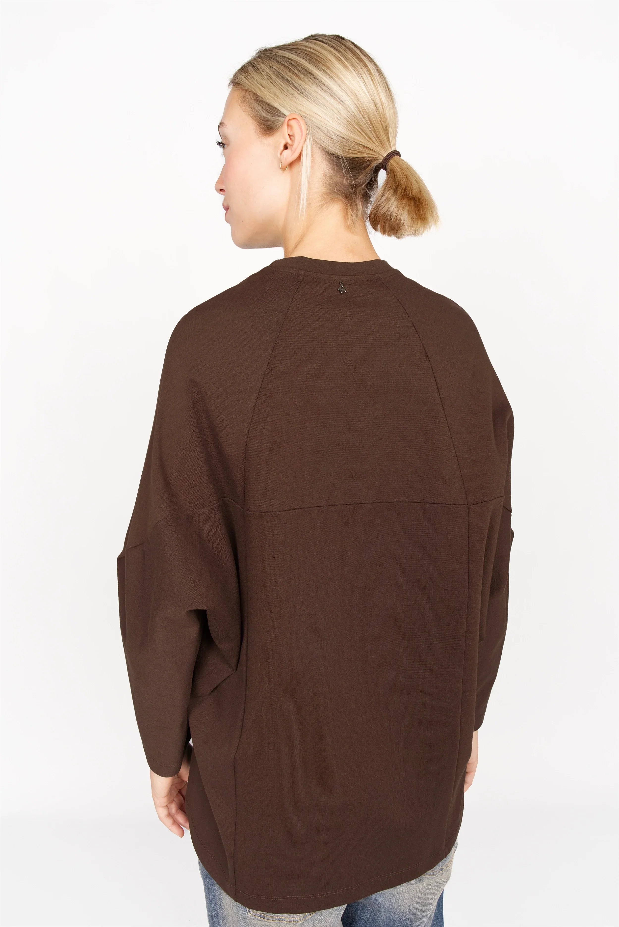 Frau trägt braunes TOOVE Shirt aus elastischem Interlock-Jersey mit verlängertem Rücken und überschnittenen Schultern, Rückansicht.