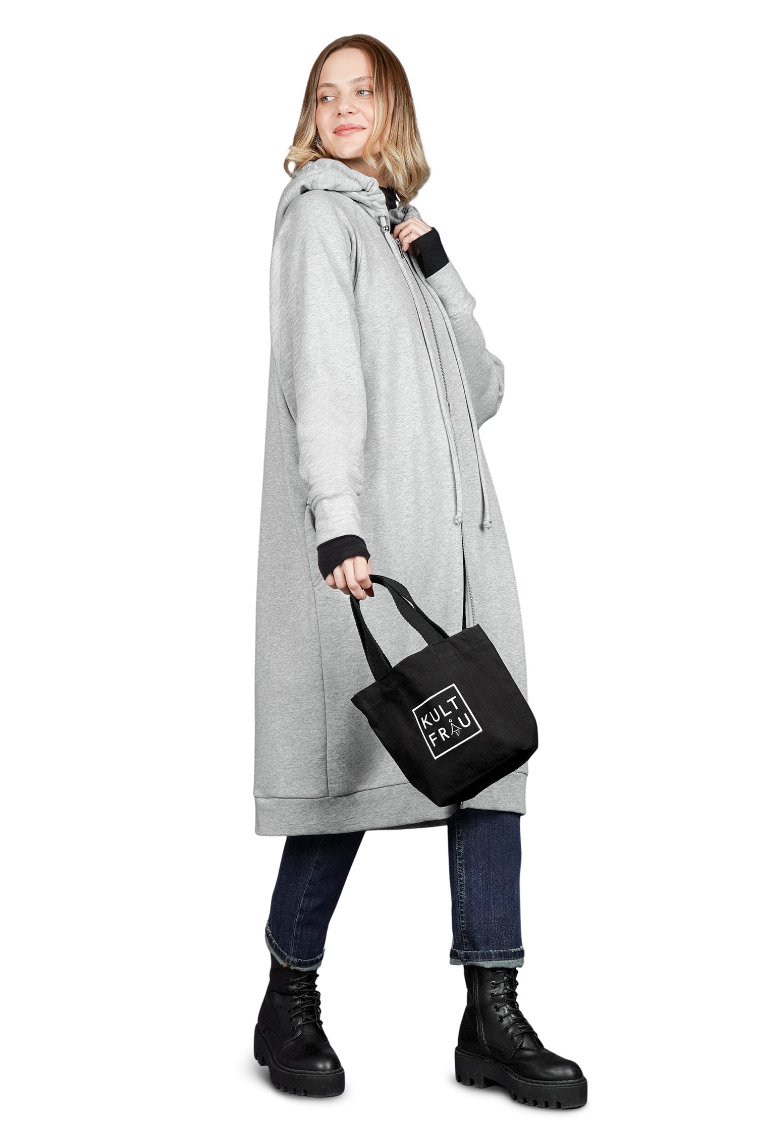 Eine Frau steht seitlich vor einem weißen Hintergrund und hält eine kleine, schwarze Stofftasche in ihrer rechten Hand. Die Tasche zeigt ein weißes, quadratisches Logo mit dem Brandnamen "KULTFRAU" in Großbuchstaben. Sie trägt einen oversized grauen Hoodie mit Kapuze und Reißverschluss, eine dunkelblaue Jeans und schwarze Schnürstiefel mit einer dicken Sohle. Ihre Haare sind mittellang und blond, und sie hat ein leichtes Lächeln. Die Tasche wirkt kompakt und minimalistisch, passend für kleinere Gegenstände