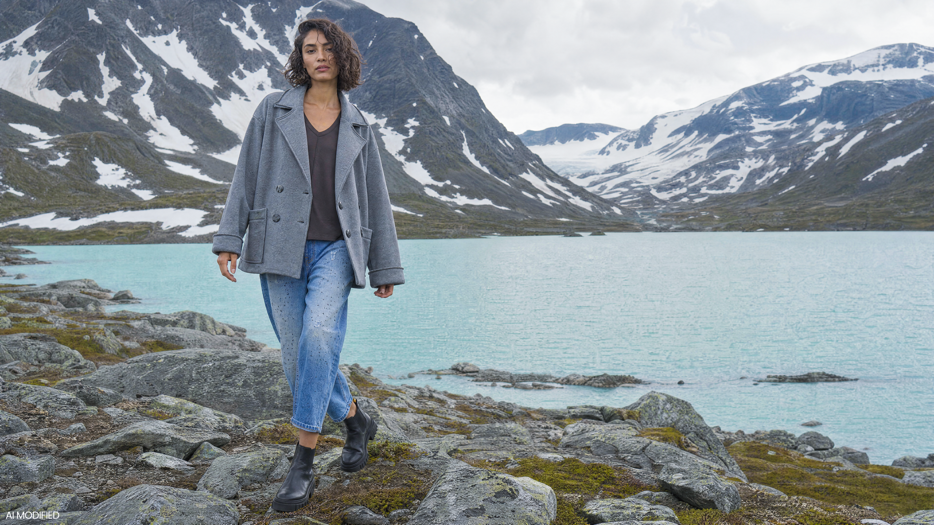 Frau in grauer Jacke und Jeans vor einem türkisfarbenen Bergsee und schneebedeckten Gipfeln.