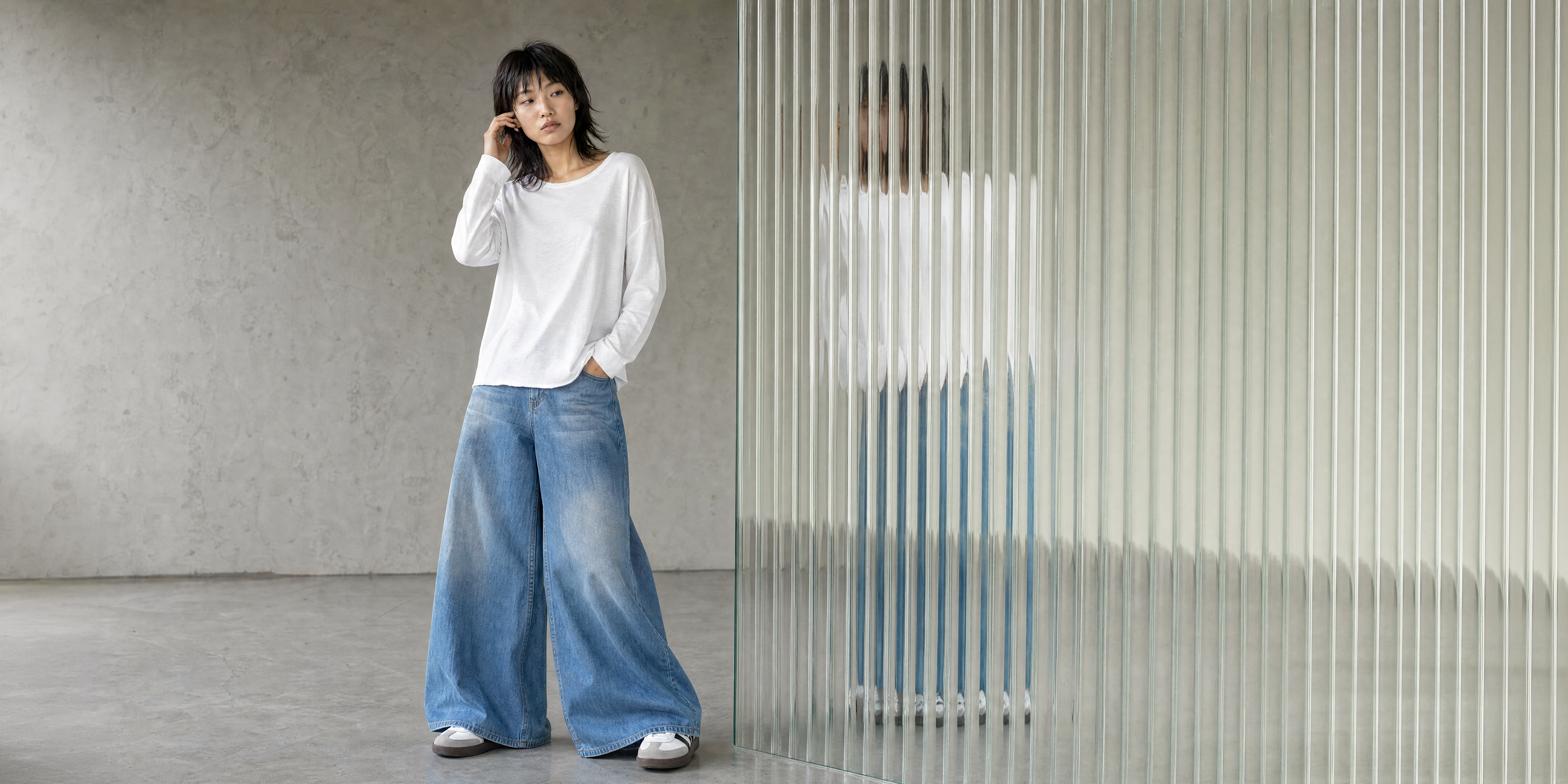 Model in weißem Langarmshirt und weiter blauer Jeans vor einer geriffelten Glaswand mit Spiegelung.
