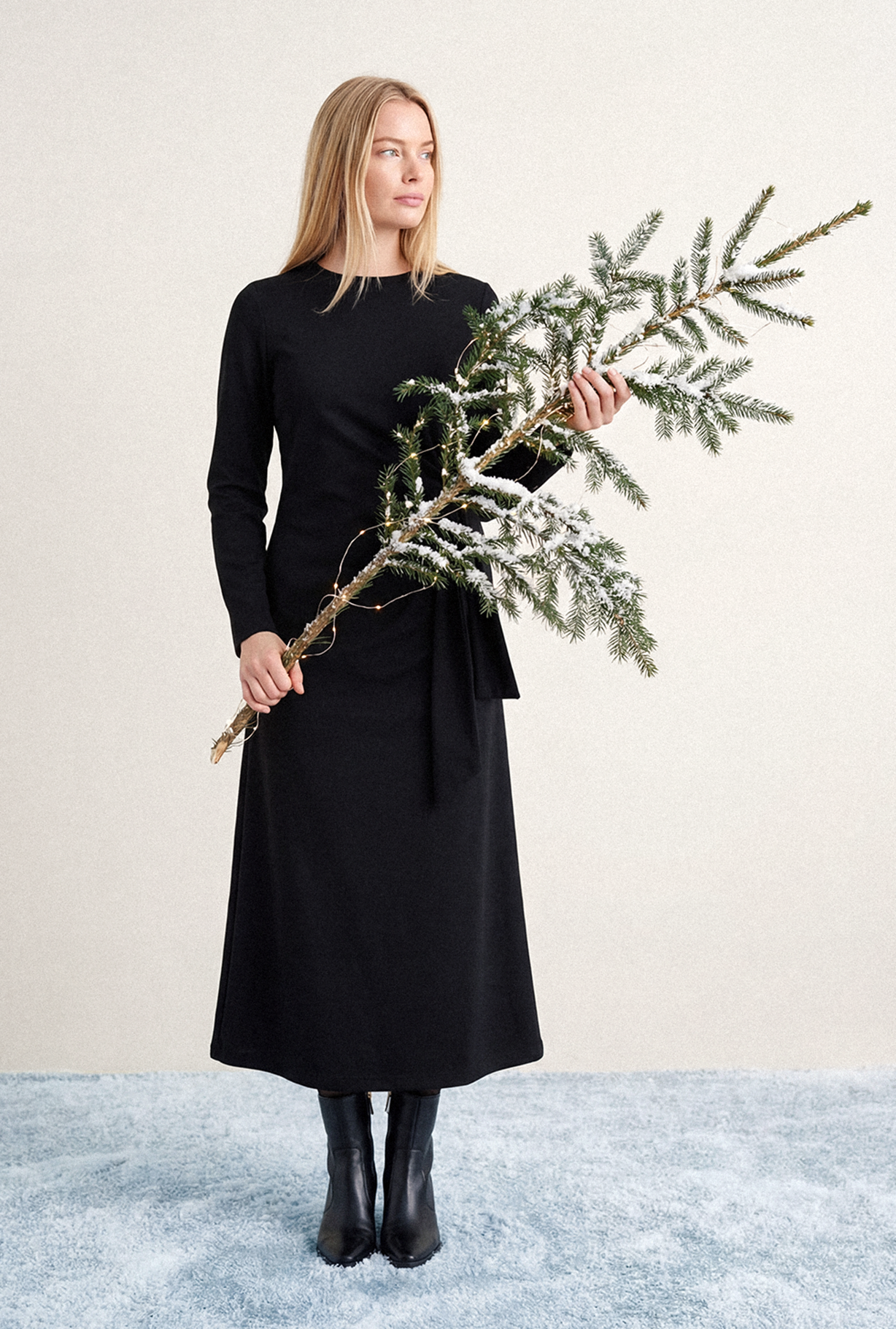 Frau in einem langen, schwarzen Kleid hält einen verschneiten Tannenzweig mit Lichterkette; sie steht auf blauem Teppich und blickt zur Seite.