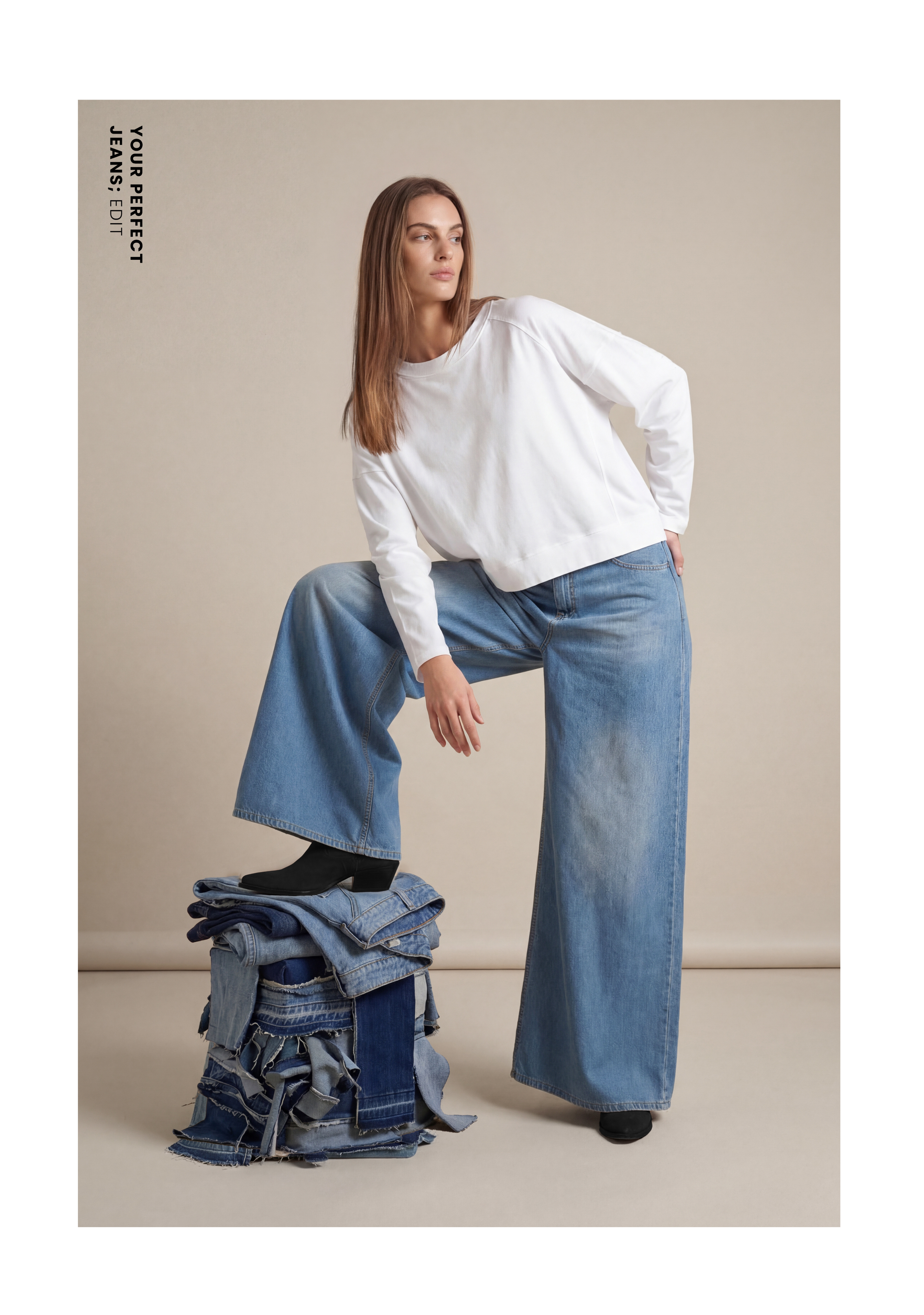 Model in lockerem weißem Longsleeve, hell ausgewaschener Wide-Leg-Jeans und schwarzen Schuhen, posiert mit einem Fuß auf einem Stapel aus gefalteten Jeans vor beigefarbenem Hintergrund.