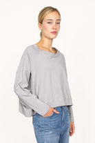 Frau trägt graues OSASU Cropped Longsleeve mit weitem Rundhalsausschnitt und locker fallender Kastenform.