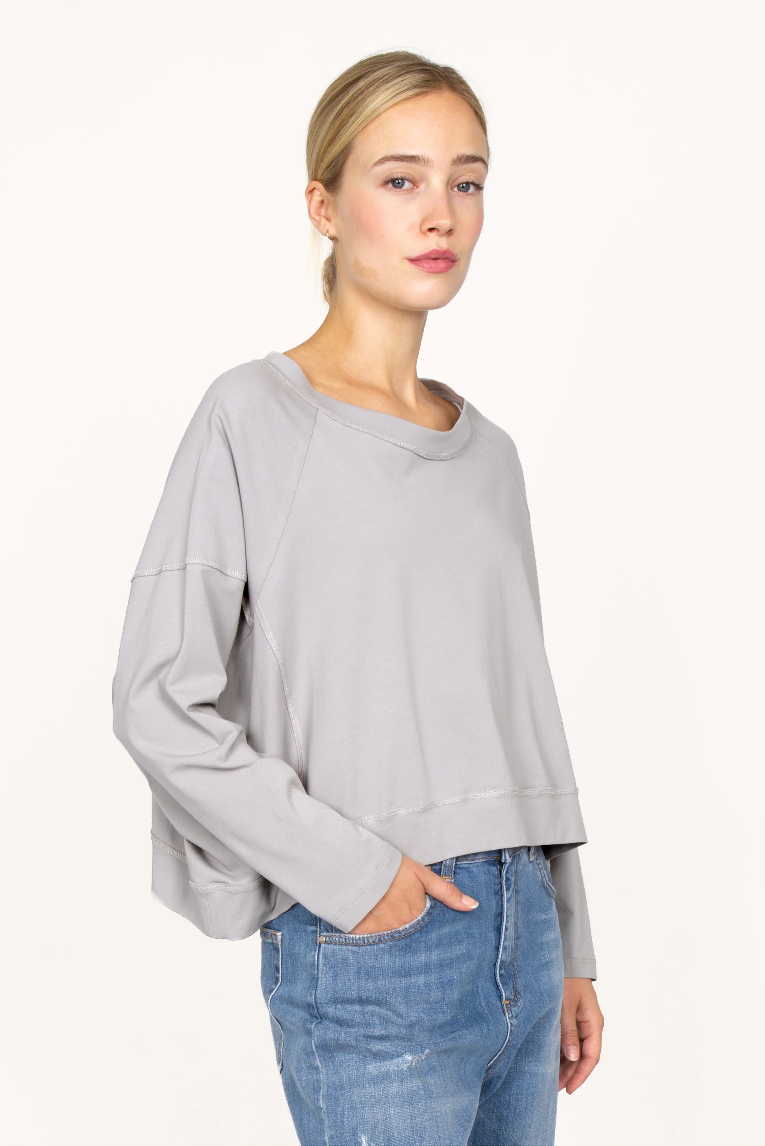 Frau trägt graues OSASU Cropped Longsleeve mit weitem Rundhalsausschnitt und locker fallender Kastenform.