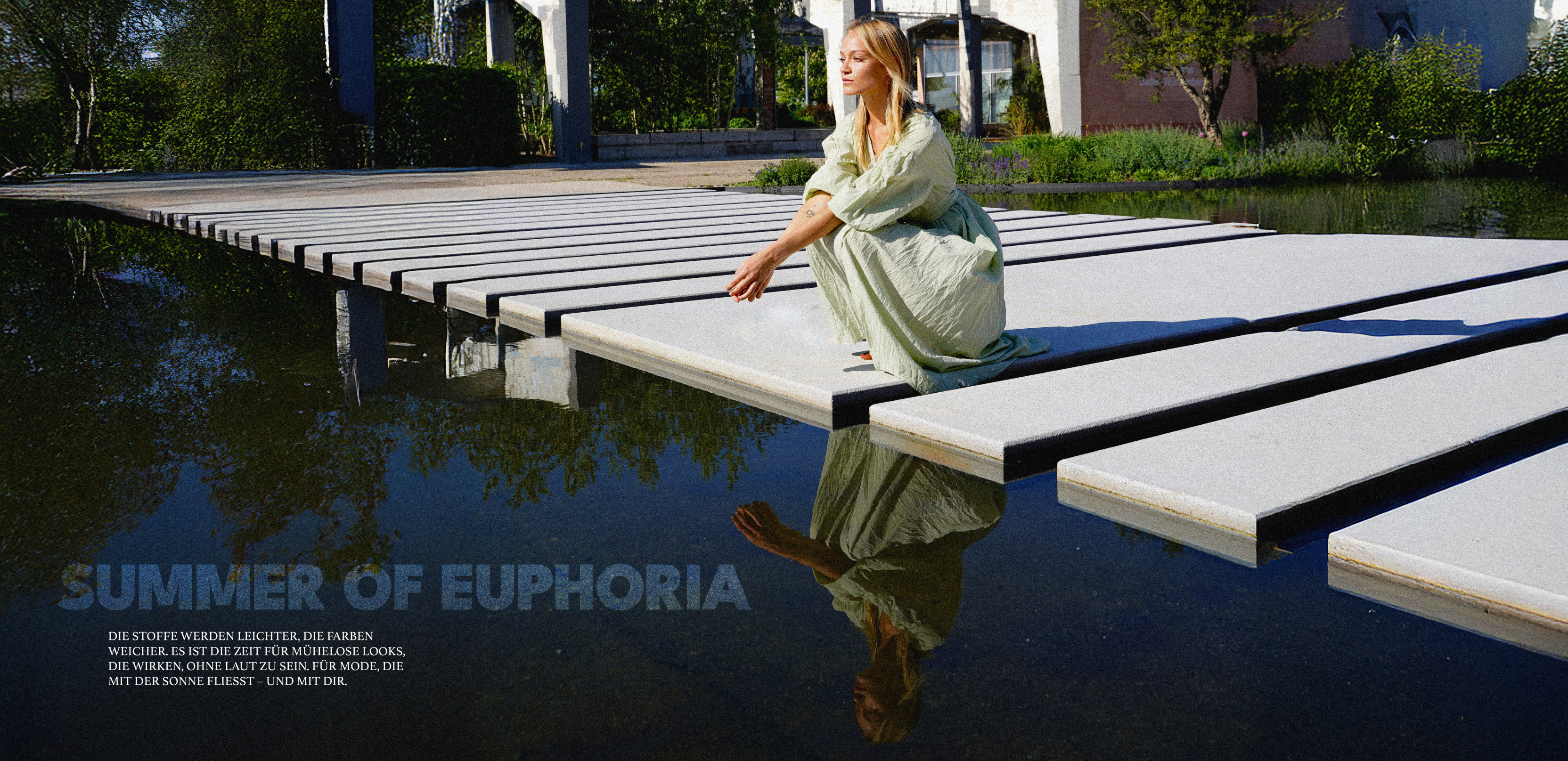 Eine Frau in einem fließenden, pastellgrünen Kleid hockt am Rand eines breiten Steg-Podestes über klarem Wasser, in dem sich ihr Spiegelbild spiegelt. Unten links ist der Schriftzug „SUMMER OF EUPHORIA“ mit weiterem deutschen Werbetext über sommerleichte Stoffe.