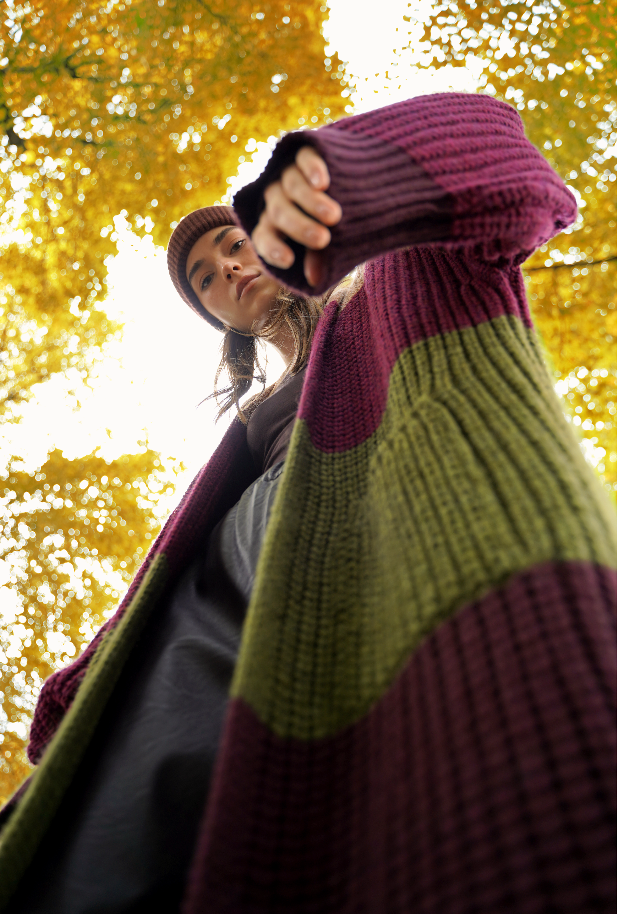 Frau trägt einen langen, grob gestrickten Mantel in Bordeaux und Olivgrün über einem dunklen Outfit und blickt aus der Froschperspektive in die Kamera, umgeben von herbstlich gelben Blättern.