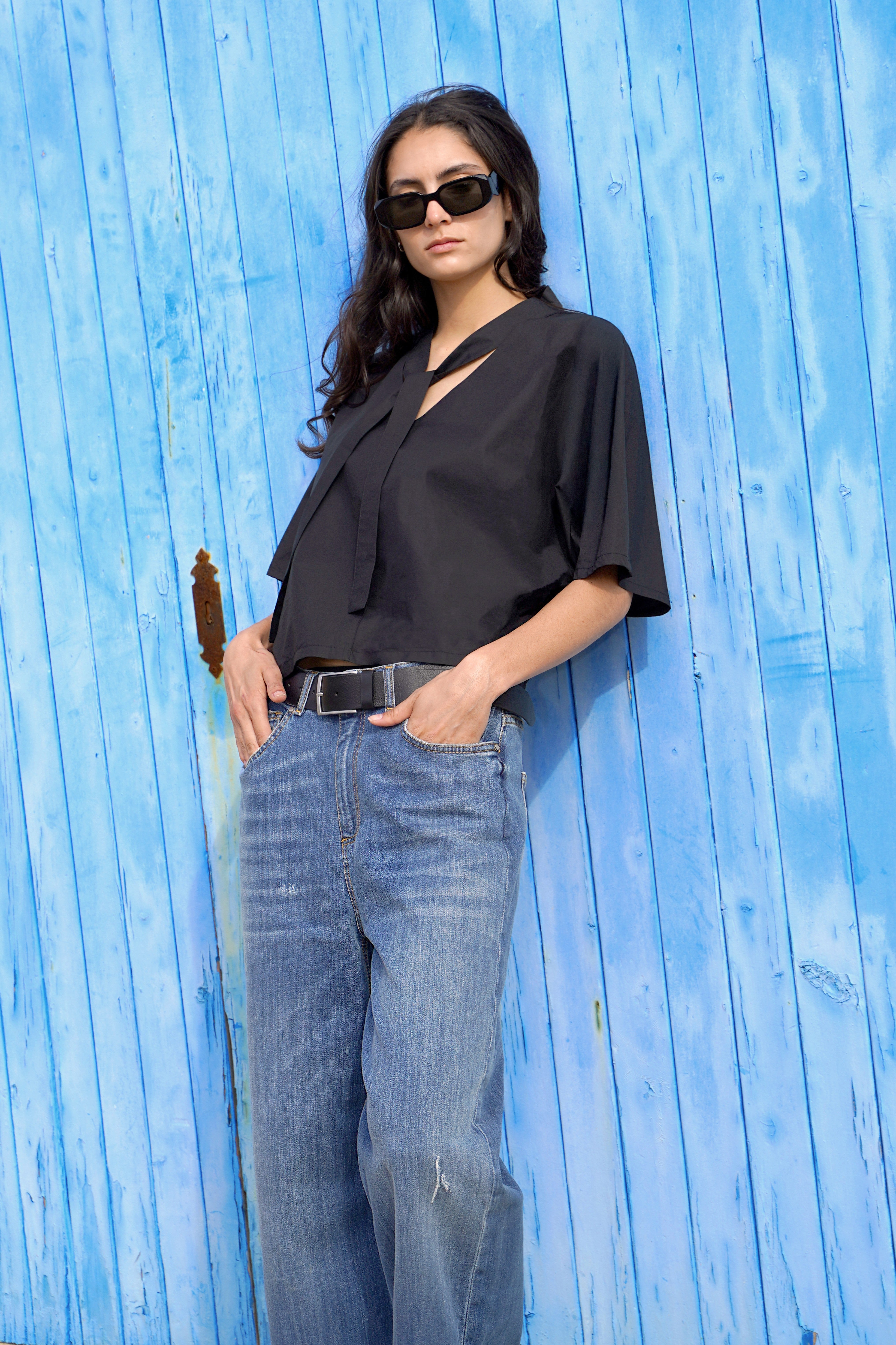 Eine Frau lehnt vor einer hellblauen, vertäfelten Holztür. Sie trägt eine schwarze, locker sitzende Bluse mit kurzem Schlipsdetail am Kragen und weite, hellblaue Jeans mit hohem Bund. Dazu kombiniert sie einen schwarzen Ledergürtel und schwarze Sonnenbrille.