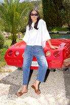 Eine Frau steht lässig an einem roten Cabrio, umgeben von Palmen und Kies. Sie trägt eine weiße, kastige Bluse mit weiten Trompetenärmeln und eine gerade geschnittene, mittlere Jeans. An den Füßen hat sie flache, cognacfarbene Lederslides und dunkle Sonnenbrille.