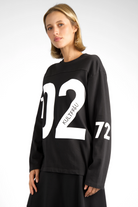 Woman wearing black AGA Statement Sweater with bold white number print on chest and sleeves in regular fit.