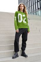 Damen trägt grünen AGA Statement Sweater mit großem weißen Zahlenprint und schwarzer Hose vor moderner Treppe