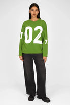 Green AGA Statement Sweater with bold white number print on front and sleeves, worn by model with black pants and shoes.