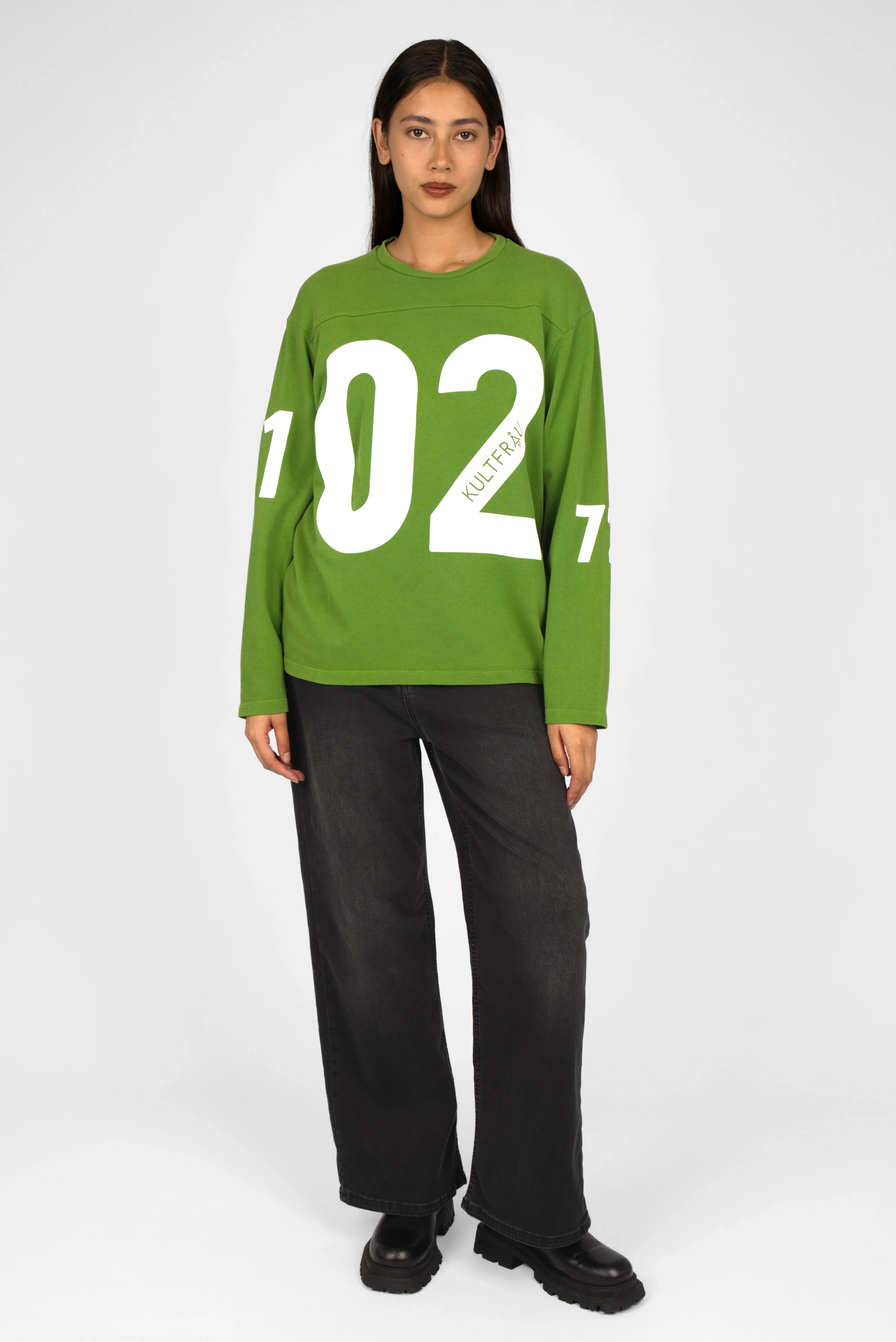 Green AGA Statement Sweater with bold white number print on front and sleeves, worn by model with black pants and shoes.
