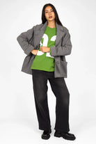 Woman wearing green AGA Statement Sweater with white number print, gray coat, and black pants standing against white background