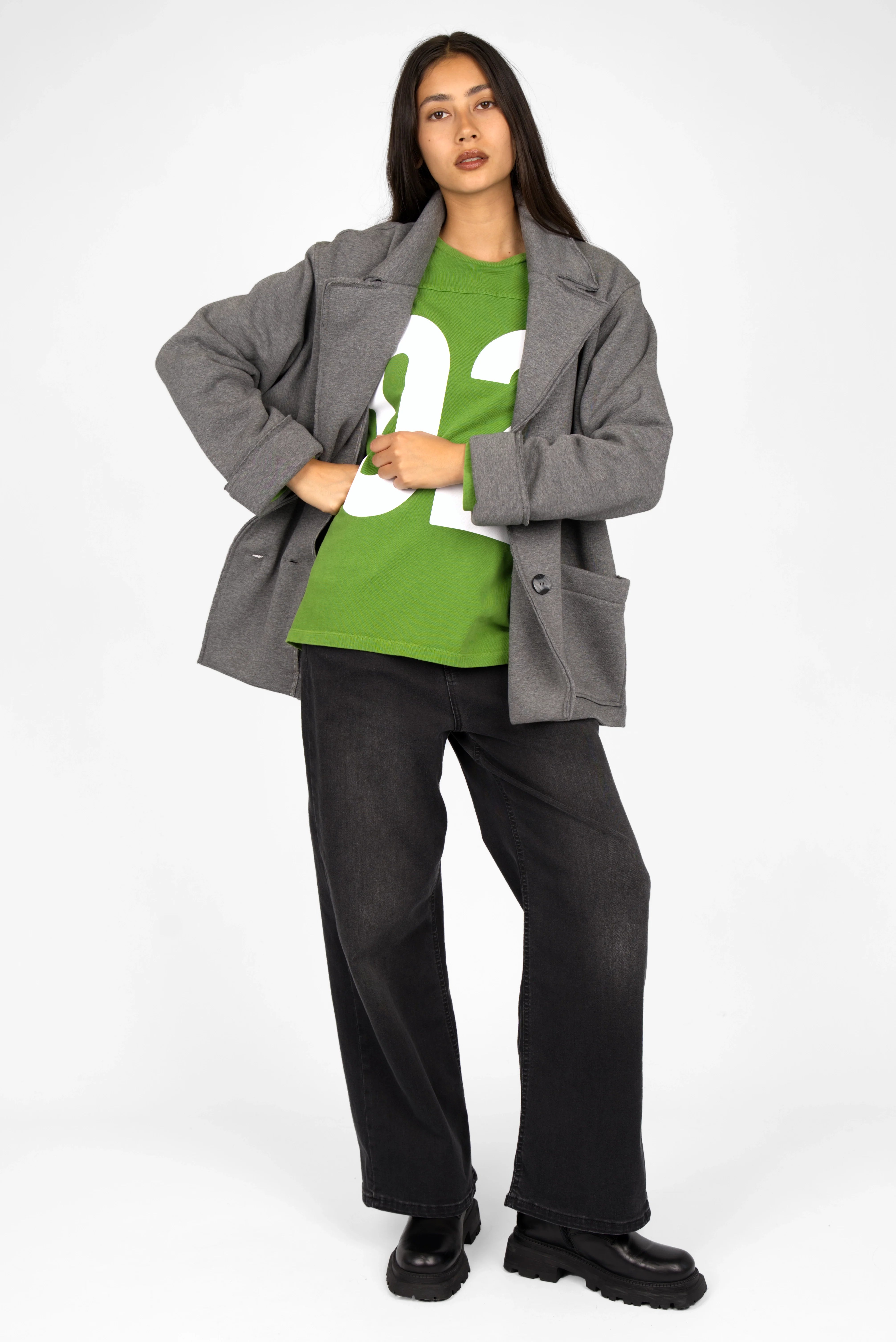 Woman wearing green AGA Statement Sweater with white number print, gray coat, and black pants standing against white background