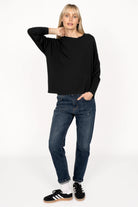 Woman wearing black ALFERA longsleeve made of lightweight viscose jersey with loose neckline and long sleeves, paired with blue jeans and black sneakers.