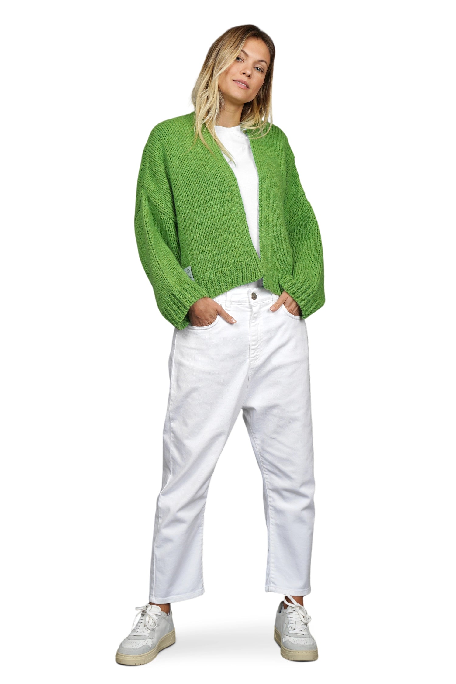 Eine junge Frau trägt eine grasgrüne, grob gestrickte Strickjacke über einem weißen T-Shirt und einer locker geschnittenen weißen Hose. Sie kombiniert das Outfit mit weißen Sneakern. Ihre Pose ist entspannt, mit leicht geneigtem Kopf und einer Hand in der Hosentasche. Der Hintergrund ist weiß.