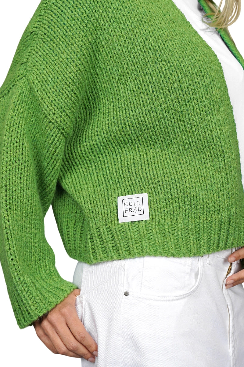 Nahaufnahme einer grasgrünen, grob gestrickten Strickjacke. Am unteren Rand ist ein weißes, quadratisches Label mit der Aufschrift „KULT FRAU“ angenäht. Die Jacke wird über einer weißen Hose getragen, eine Hand steckt locker in der Hosentasche. Der Hintergrund ist weiß.