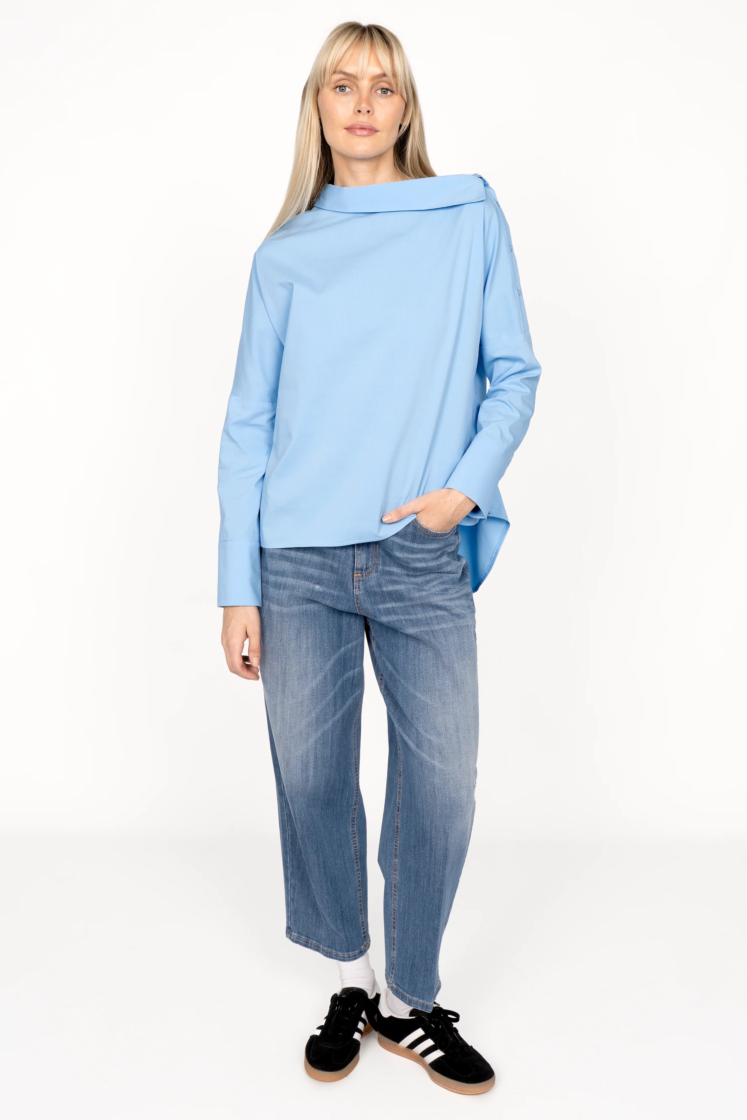 Woman wearing light blue BONI asymmetrische one shoulder Bluse with asymmetrical collar, wide cuffs, and relaxed fit paired with jeans and black sneakers.