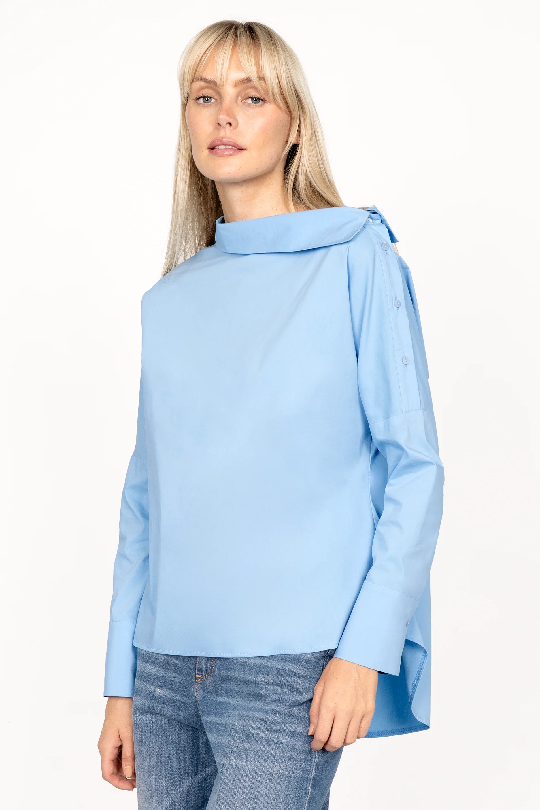 Woman wearing light blue BONI asymmetrical one shoulder blouse with extended back and buttoned wide cuffs