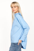 Woman wearing BONI asymmetrical one shoulder blouse in light blue with buttoned shoulder detail and extended back hem