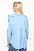 Woman wearing light blue BONI asymmetrical one shoulder blouse with long sleeves and extended back, viewed from behind.