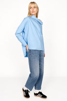 Woman wearing BONI asymmetrical one shoulder blouse in light blue with extended back and relaxed fit paired with denim jeans and black sneakers
