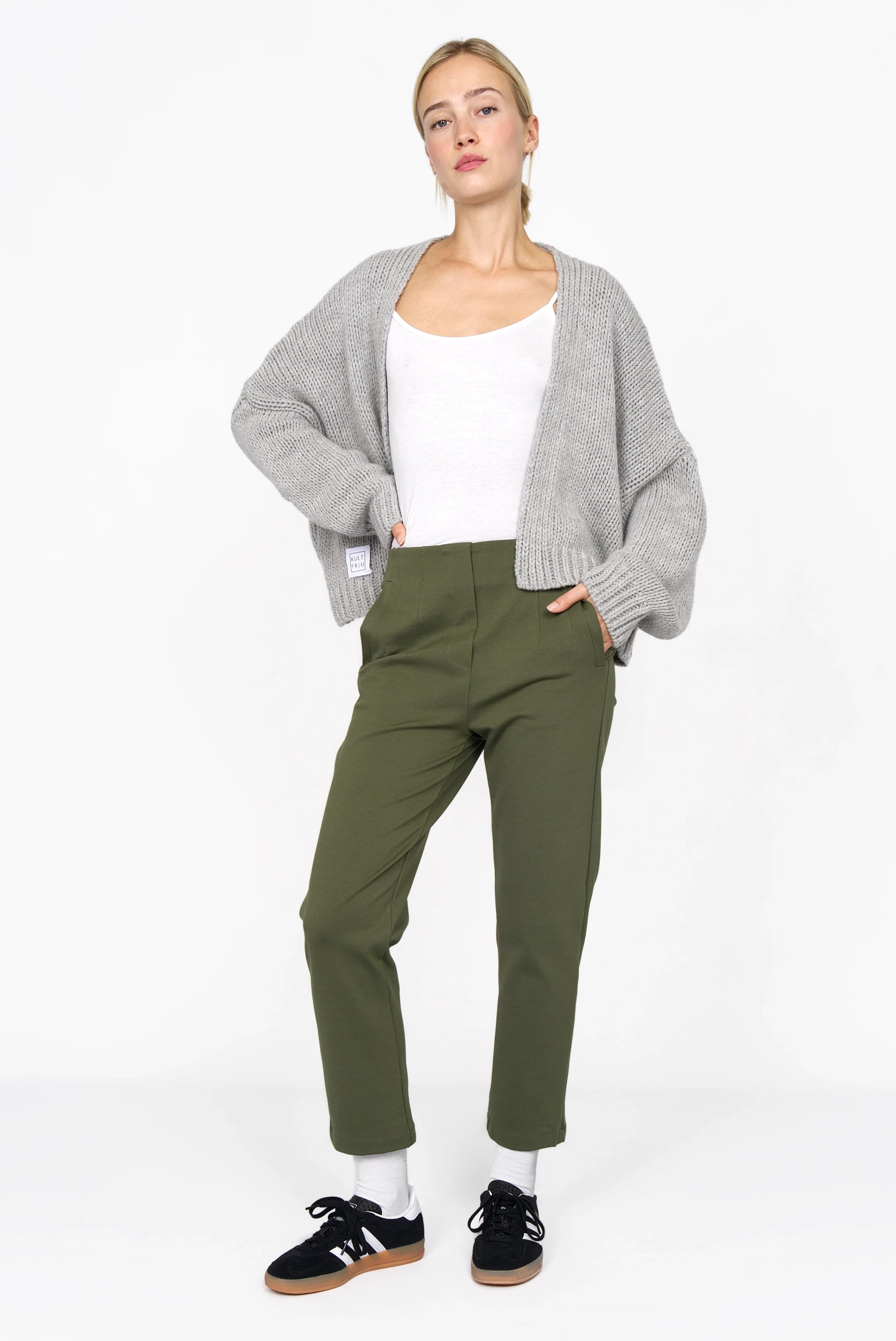 Woman wearing olive green slim fit cropped ankle pants with elastic back waist, paired with white top, gray cardigan, white socks, and black sneakers