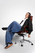 Frau sitzt entspannt im Drehstuhl trägt schwarze Relaxed Fit Sweatjacke mit Knopfleiste, weiße Bluse, blaue Jeans und schwarze Sneaker