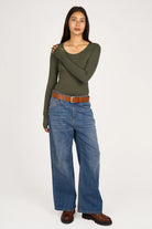 Woman wearing olive green VALUM Longsleeve with U-boat neckline, extra long sleeves with thumb holes, paired with blue wide-leg jeans and brown belt.