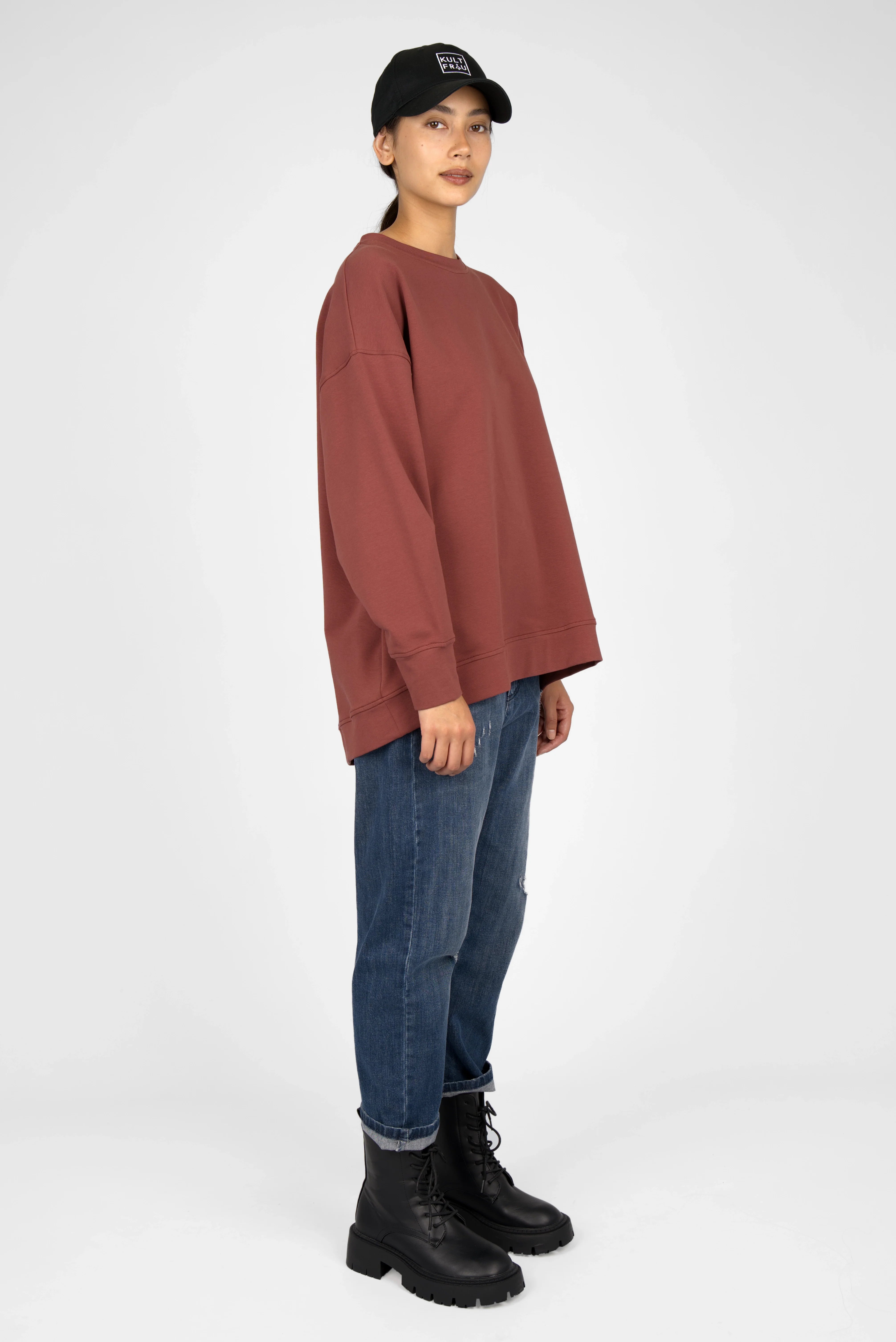 Woman wearing a rust-colored EIREL oversize sweater with dropped shoulders and relaxed fit paired with blue jeans and black boots