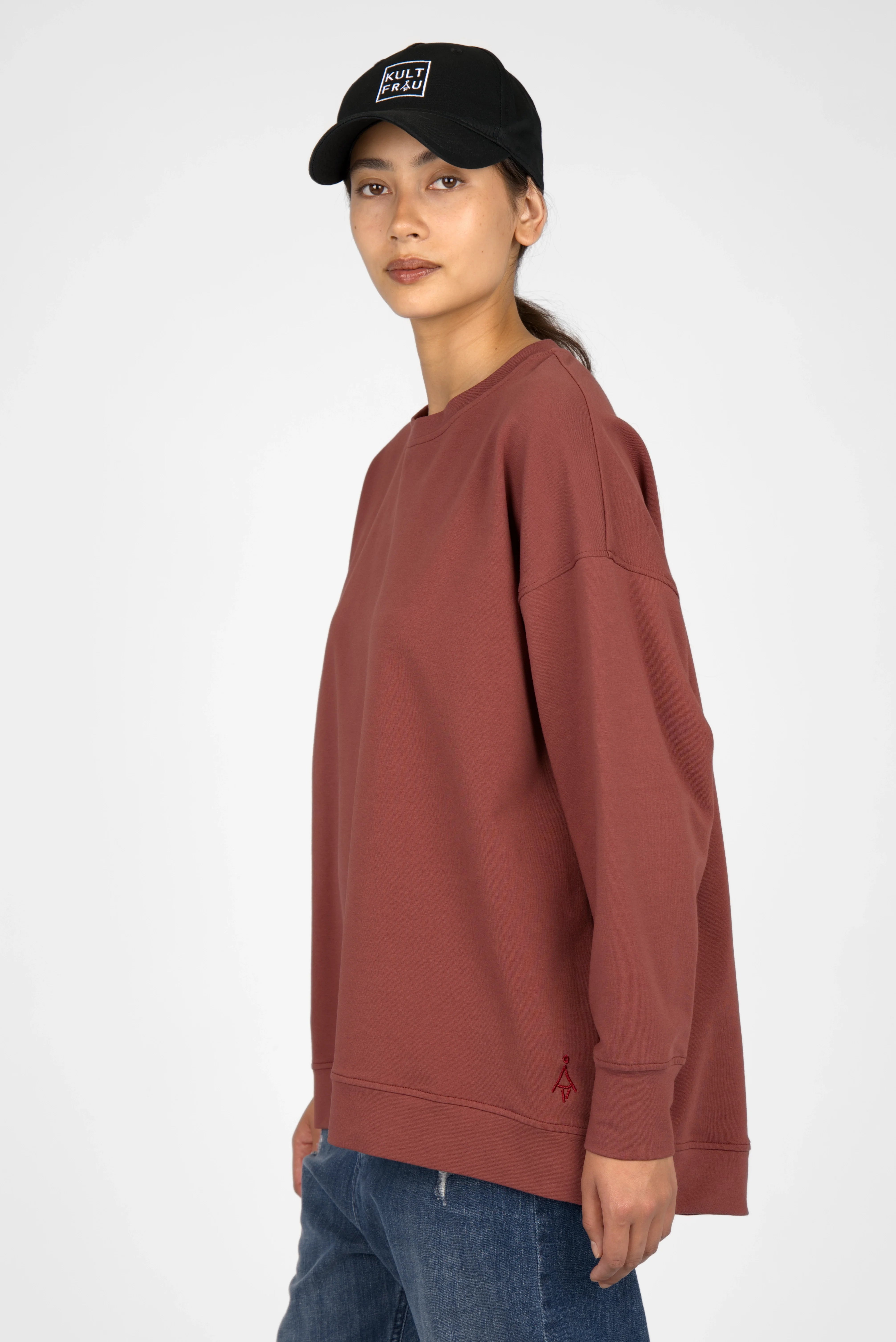 Woman wearing a brown EIREL oversize sweater with dropped shoulders and tonal logo embroidery, paired with blue jeans and a black cap