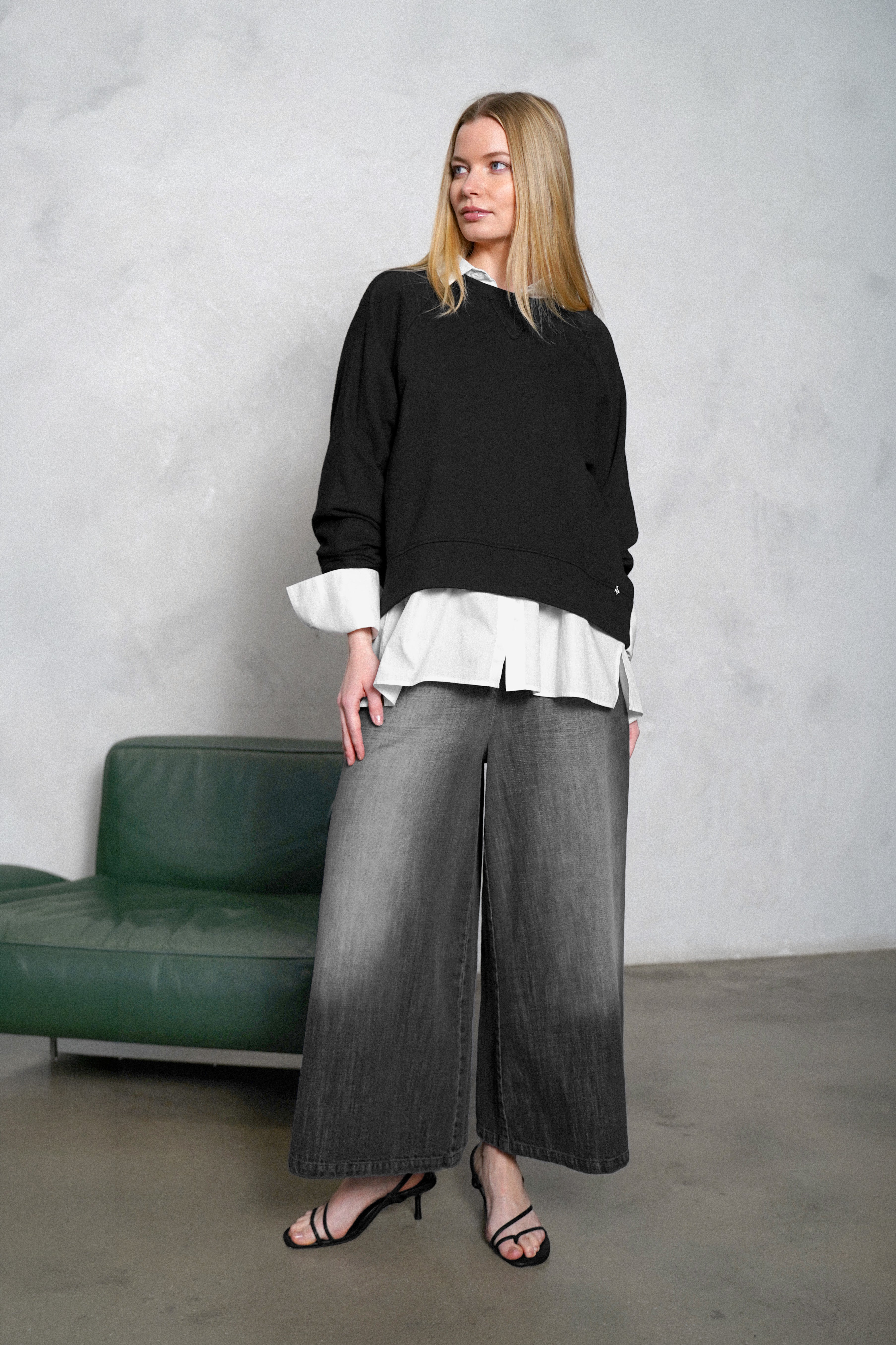 Model steht vor grauer Wand neben grünem Sofa und trägt ein schwarzes Sweatshirt über weißer Hemdbluse, dazu eine graue Wide-Leg-Jeans und schwarze Sandaletten.