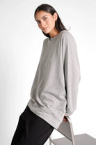 Woman wearing grey oversized GRACEY.K Sweater with round neckline and tonal logo embroidery sitting on chair
