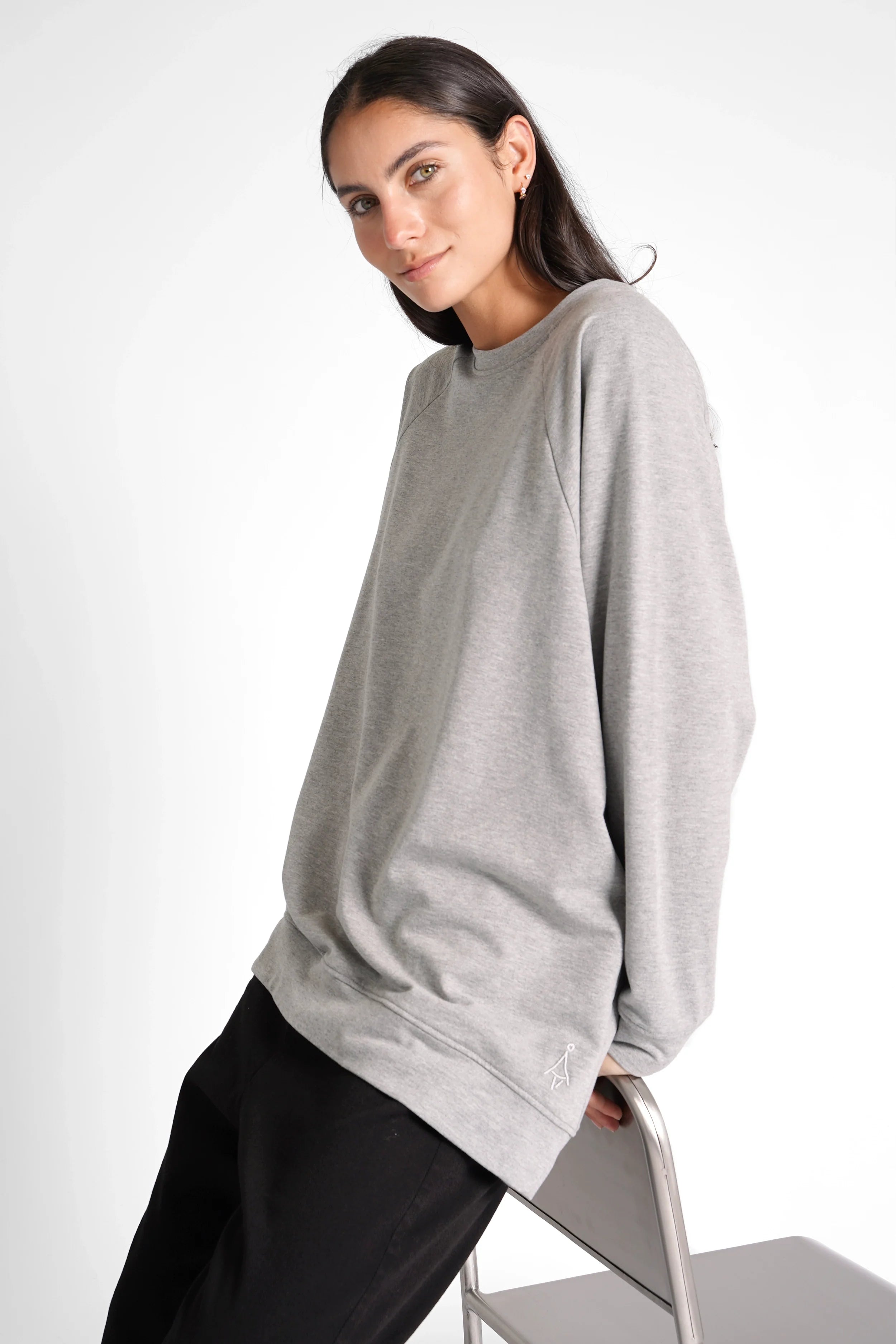 Woman wearing grey oversized GRACEY.K Sweater with round neckline and tonal logo embroidery sitting on chair