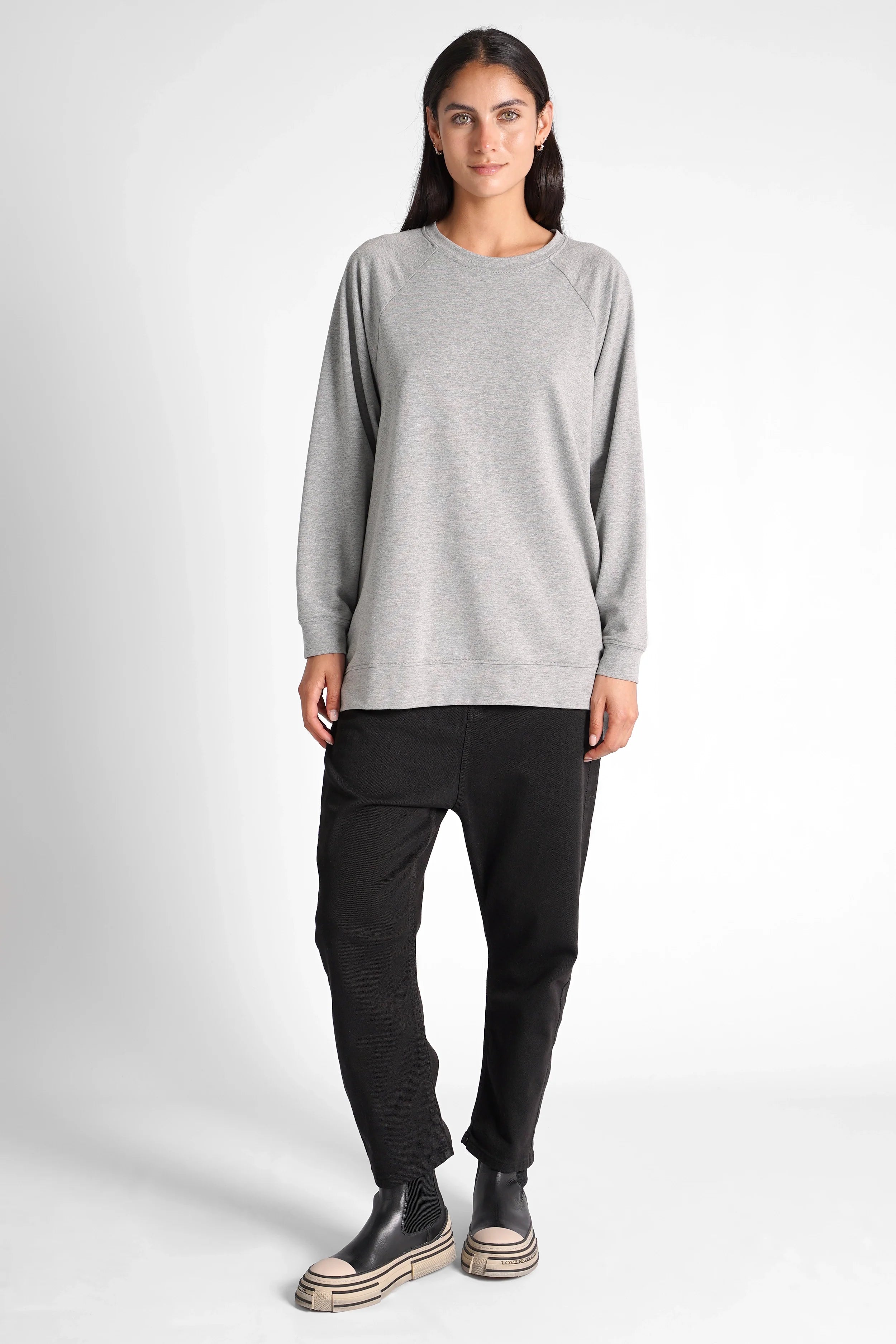 Woman wearing a grey oversized GRACEY.K Sweater with rounded hem and tonal logo embroidery, paired with black pants and black platform shoes.