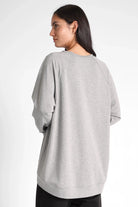 Woman wearing a grey oversized GRACEY.K Sweater with A-line and longer back hem, soft fabric and tonal logo embroidery.