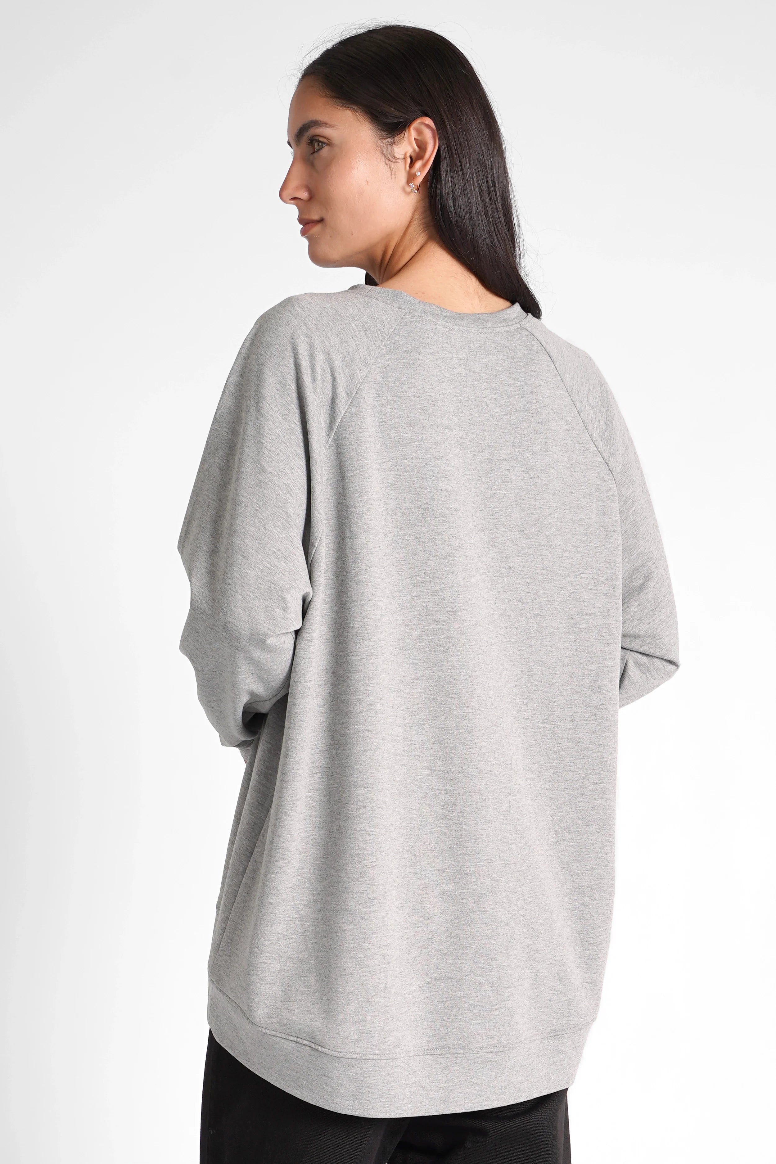 Woman wearing a grey oversized GRACEY.K Sweater with A-line and longer back hem, soft fabric and tonal logo embroidery.