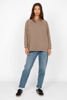 Woman wearing taupe oversized GRACEY.K Sweater with rounded hem and back slightly longer, paired with blue jeans and black sneakers.