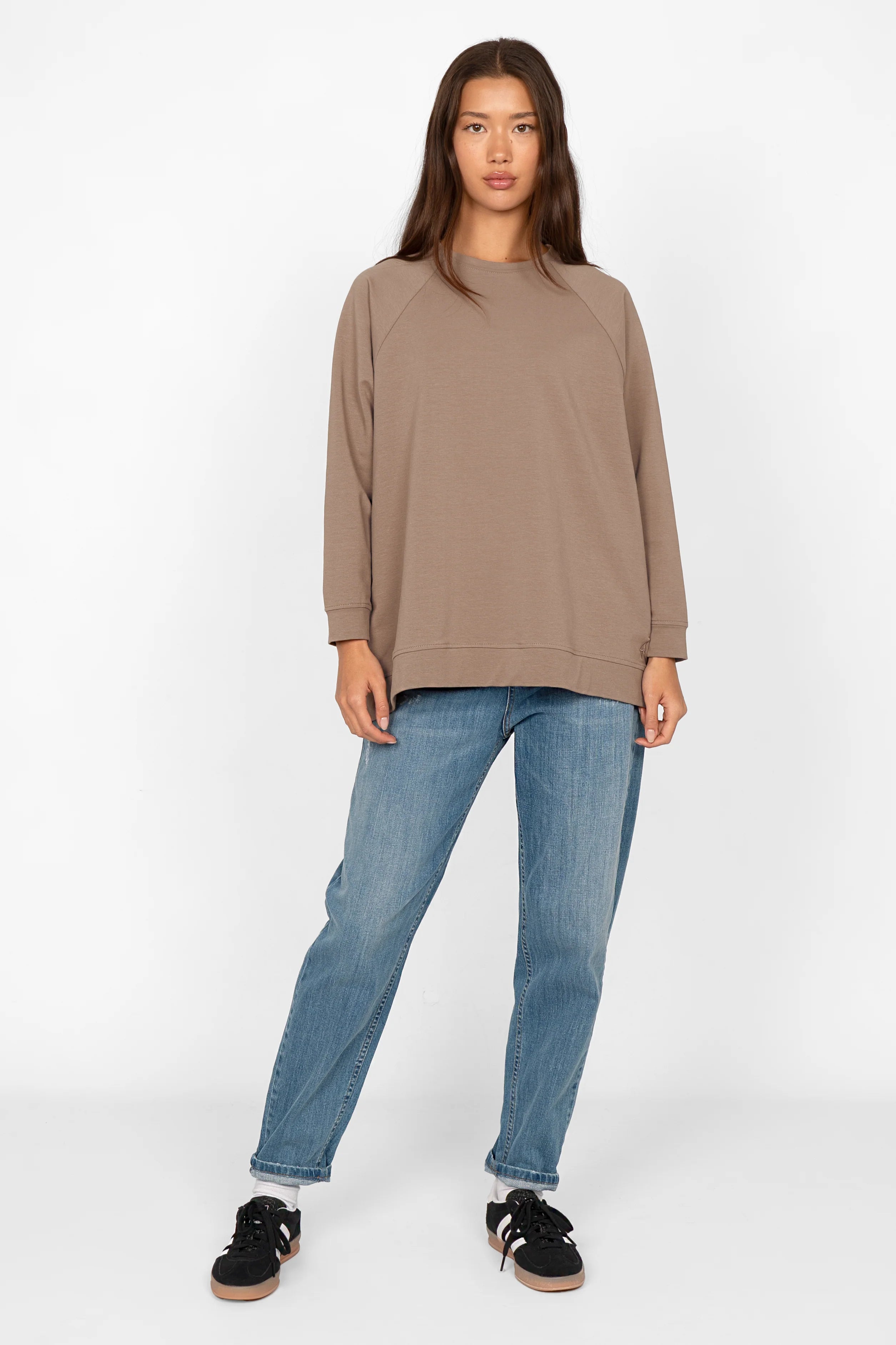 Woman wearing taupe oversized GRACEY.K Sweater with rounded hem and back slightly longer, paired with blue jeans and black sneakers.