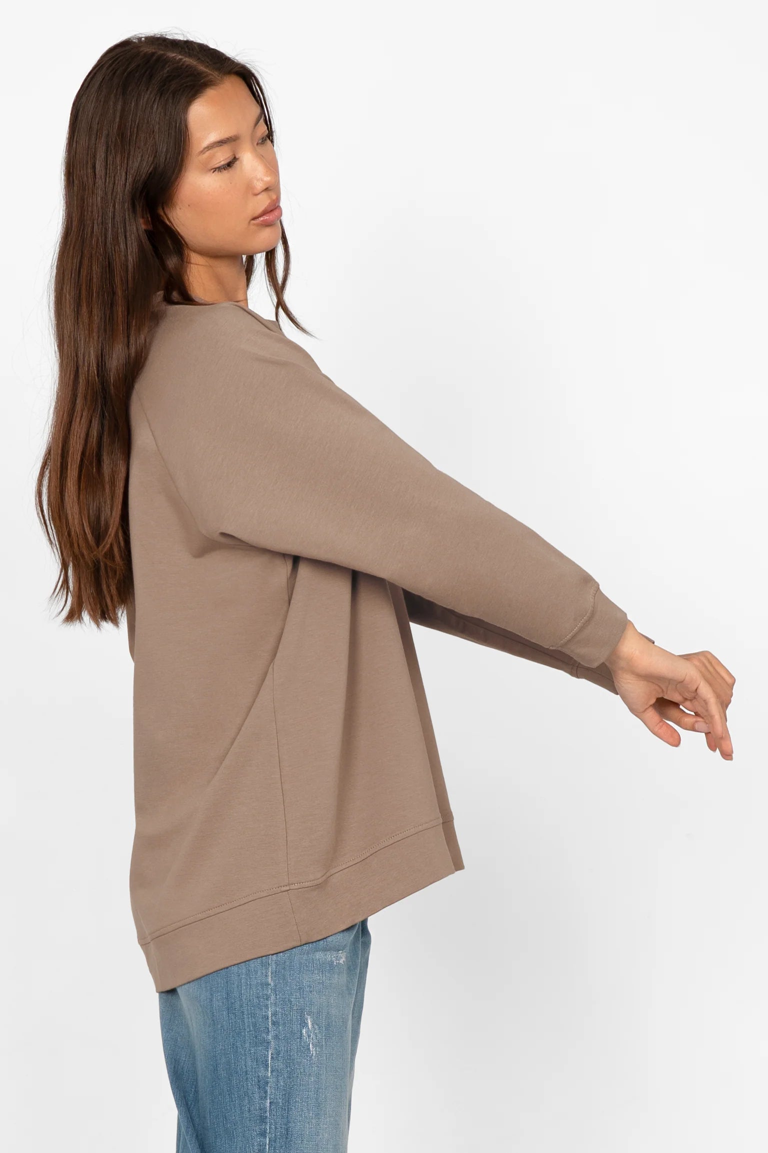 Woman wearing taupe GRACEY.K Sweater with oversized fit and rounded hem, paired with blue jeans, side view