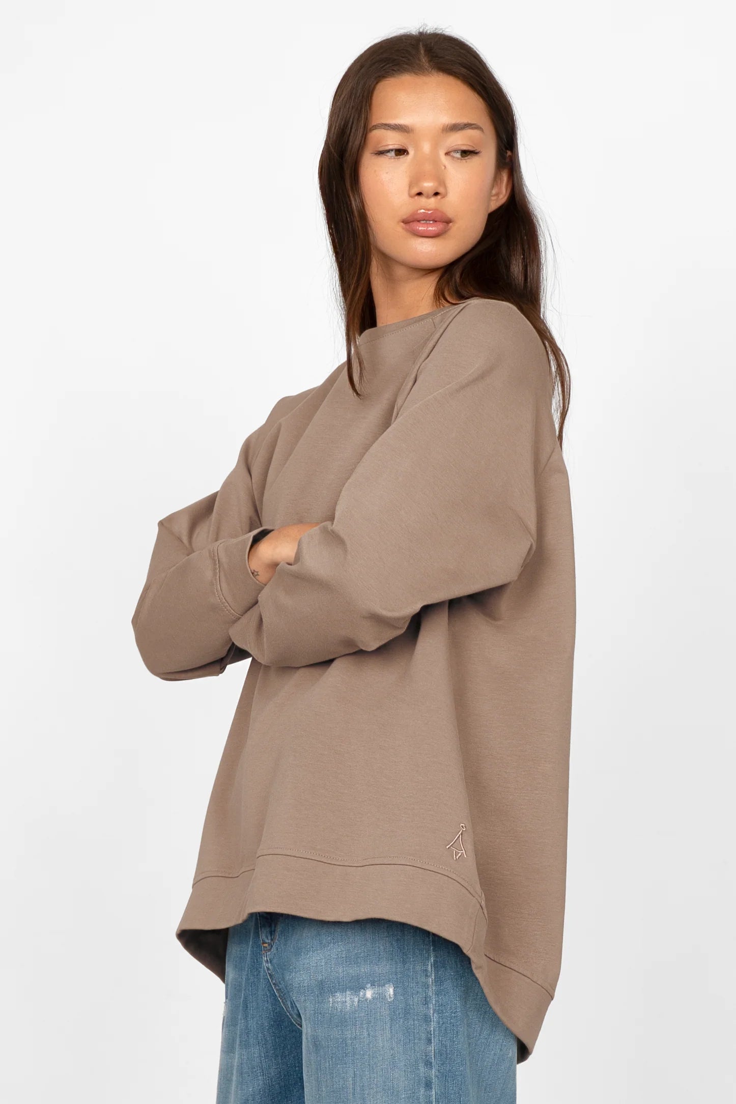 Woman wearing taupe oversized GRACEY.K Sweater with rounded hem and tonal logo embroidery, paired with blue jeans.
