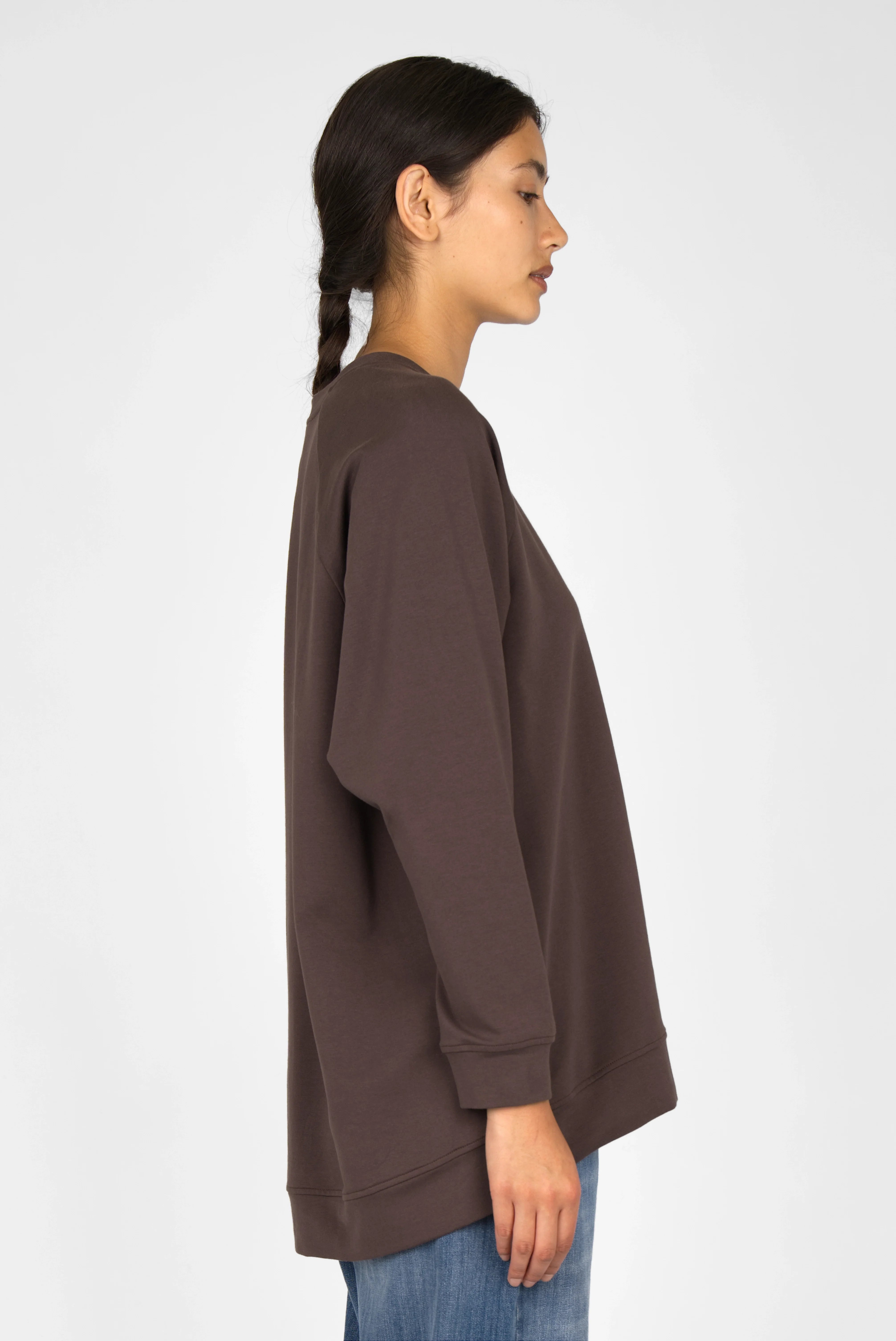Side view of a woman wearing a brown GRACEY.K Sweater with oversized fit, round neckline, and longer back hem.
