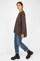 Woman wearing oversized brown GRACEY.K Sweater with rounded hem and tonal logo embroidery, paired with blue jeans and black boots.