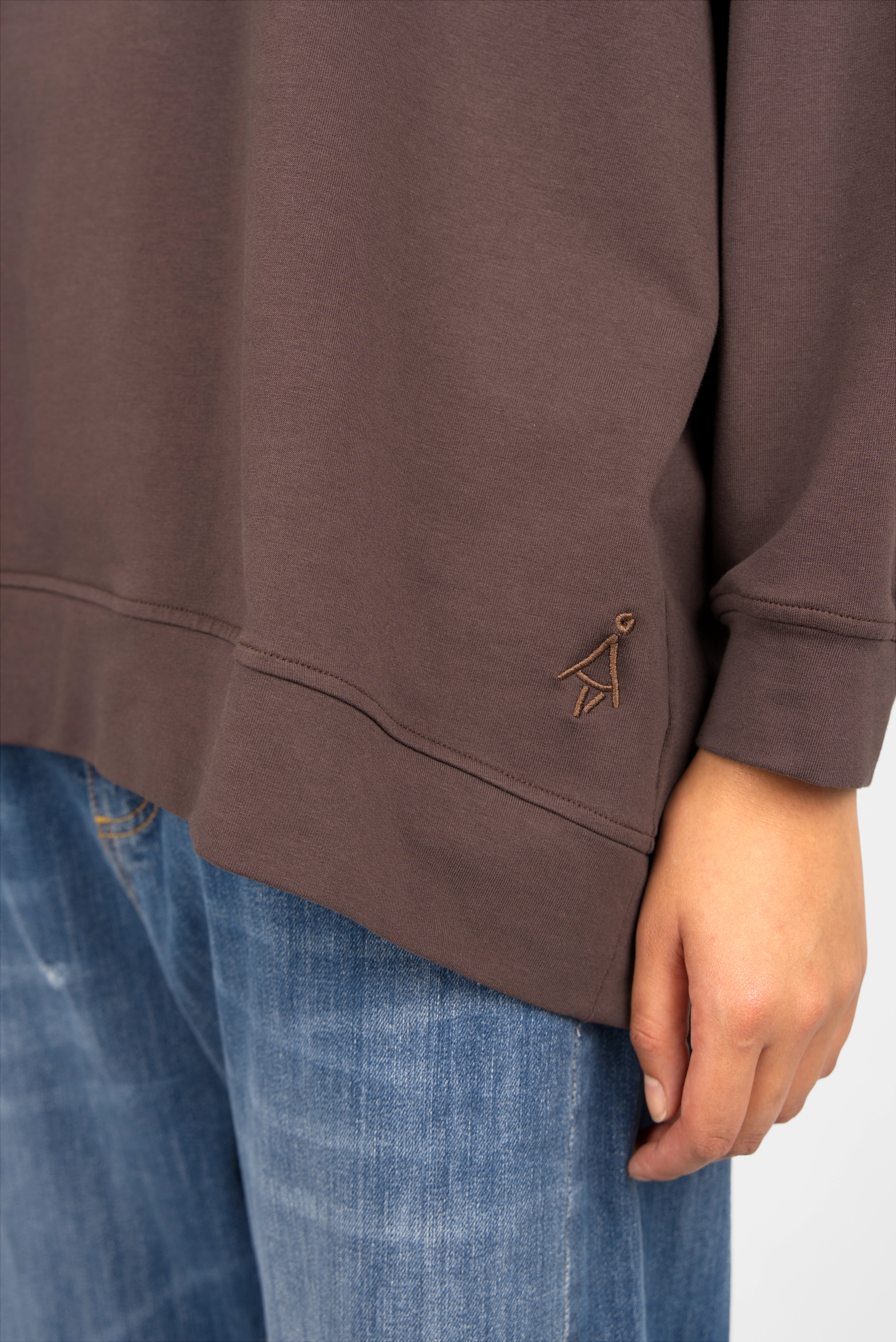 Close-up of a brown GRACEY.K Sweater with tonal logo embroidery and rounded hem on a person wearing blue jeans