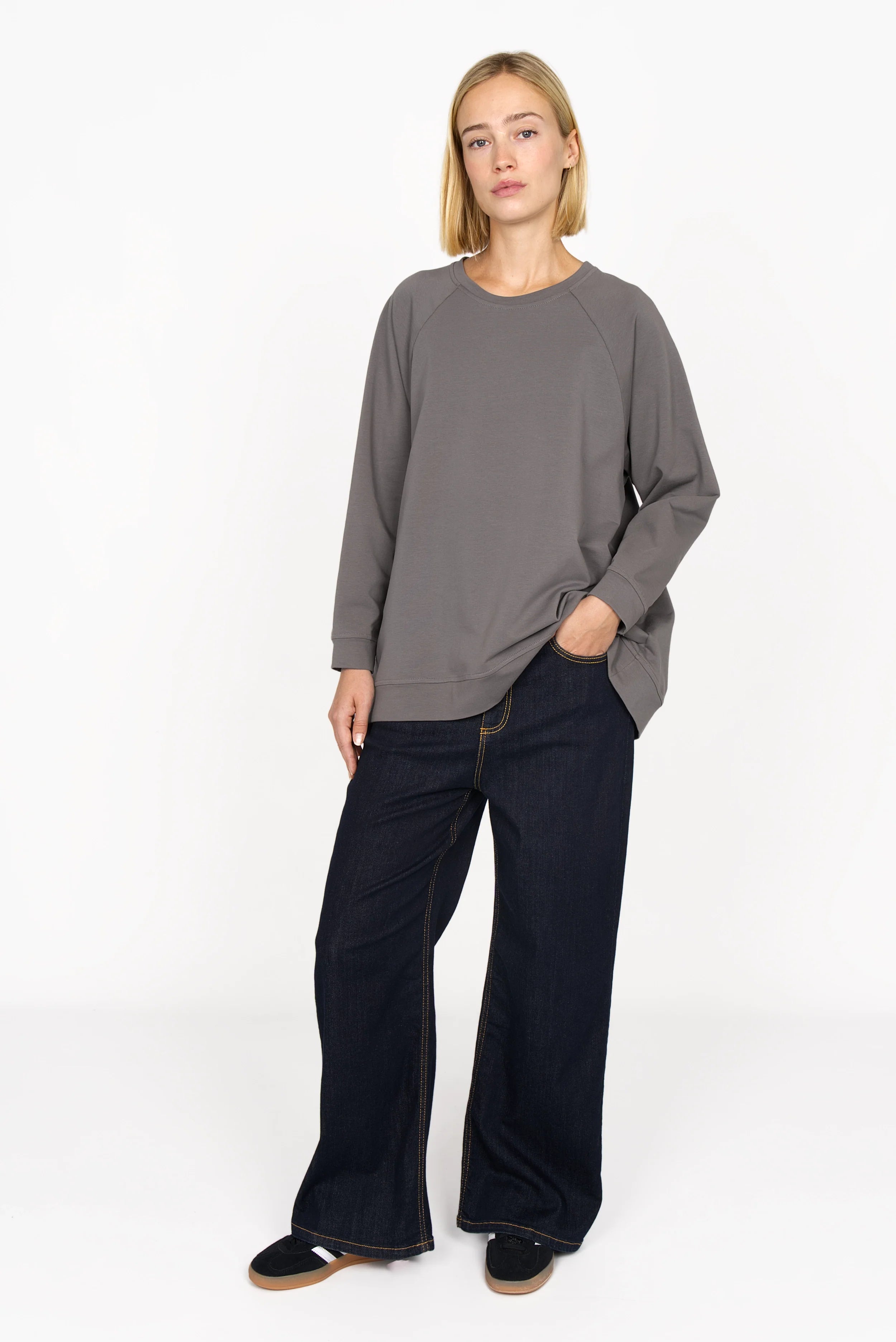 Woman wearing gray GRACEY.K Sweater with round neckline and tonal logo embroidery, paired with wide-leg dark jeans.