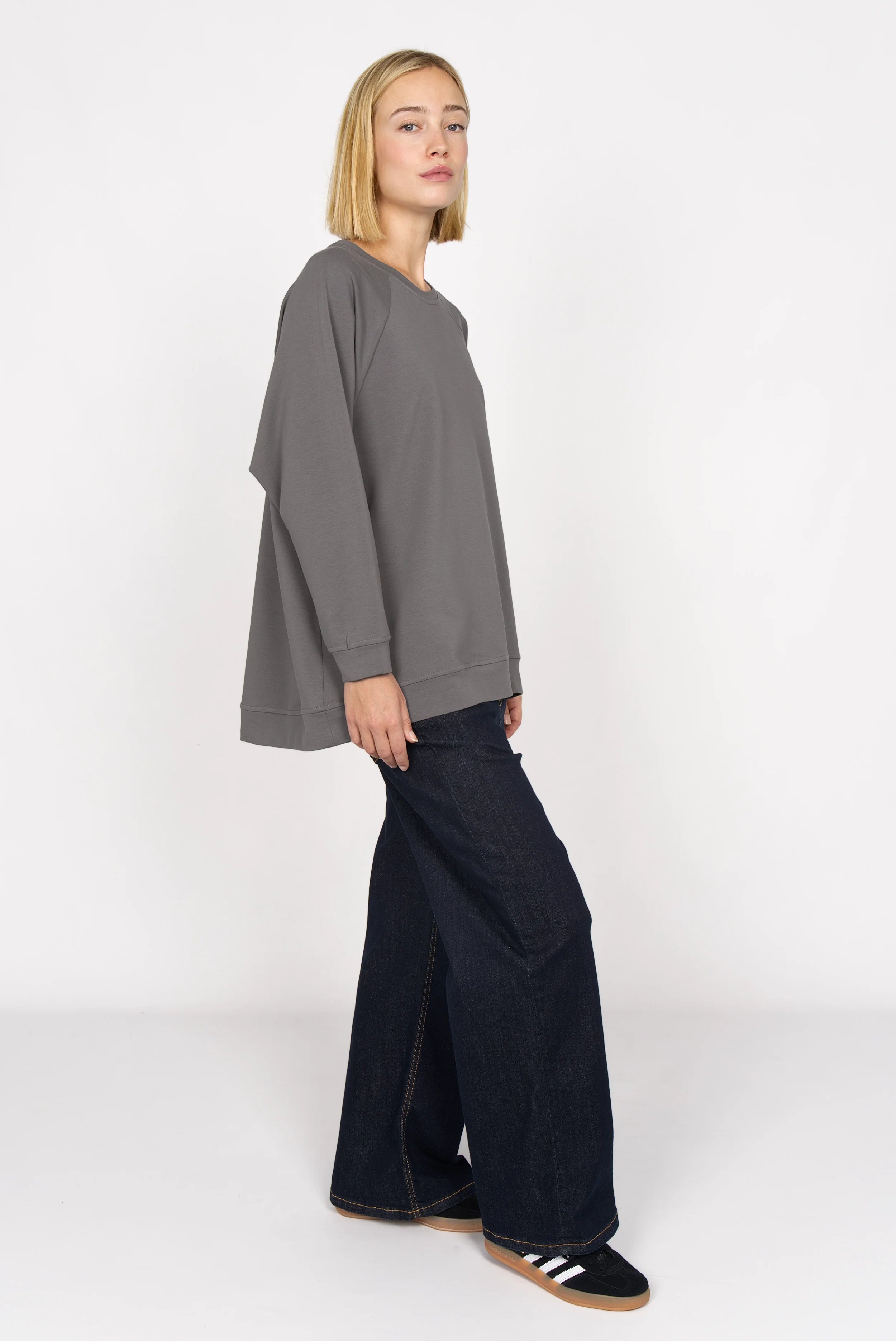 Woman wearing a gray GRACEY.K Sweater with A-line silhouette and longer back paired with wide-leg dark jeans and black sneakers.