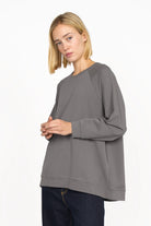 Woman wearing a gray GRACEY.K Sweater with round neckline, oversized fit, tonal logo embroidery, and longer back hem