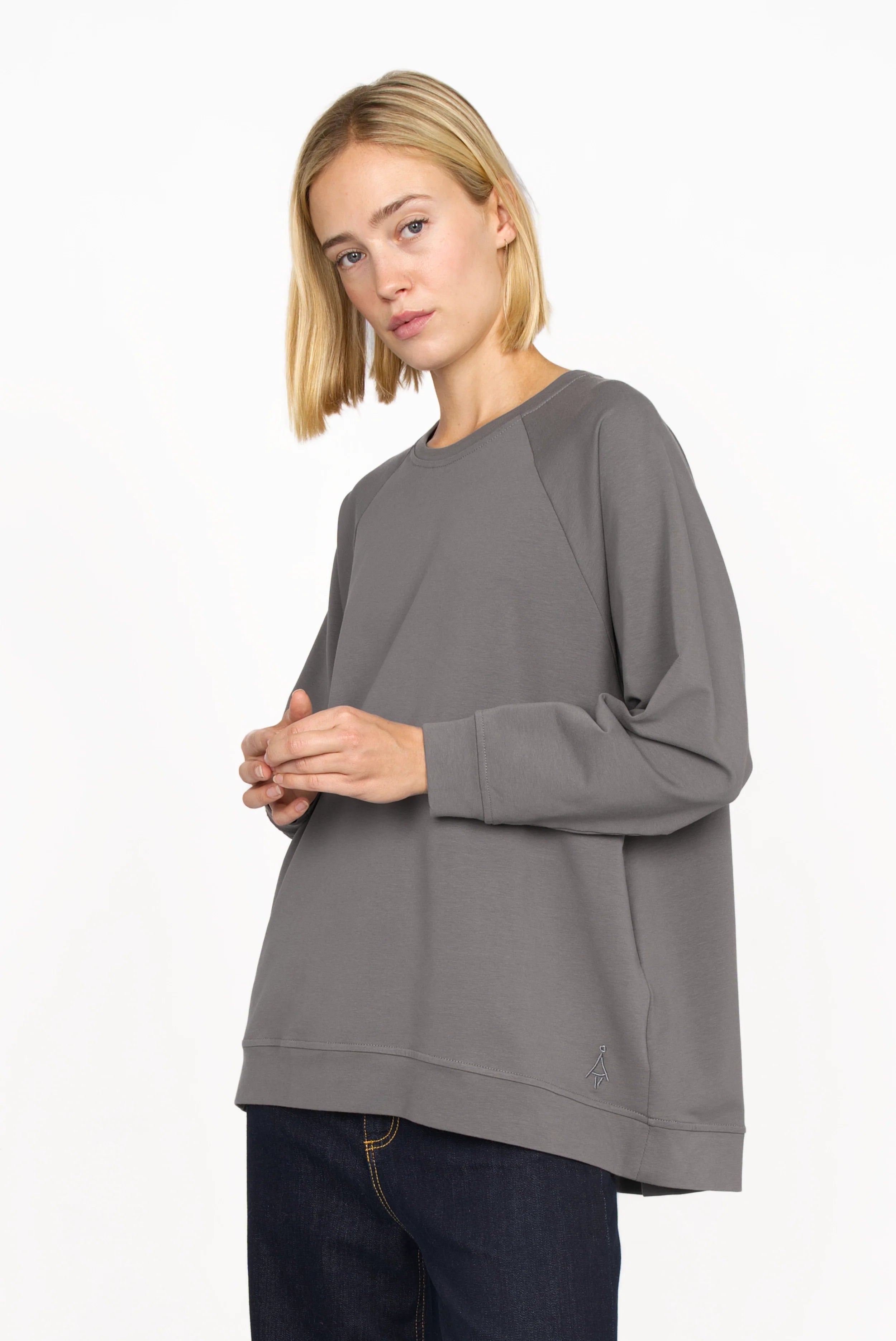 Woman wearing a gray GRACEY.K Sweater with round neckline, oversized fit, tonal logo embroidery, and longer back hem