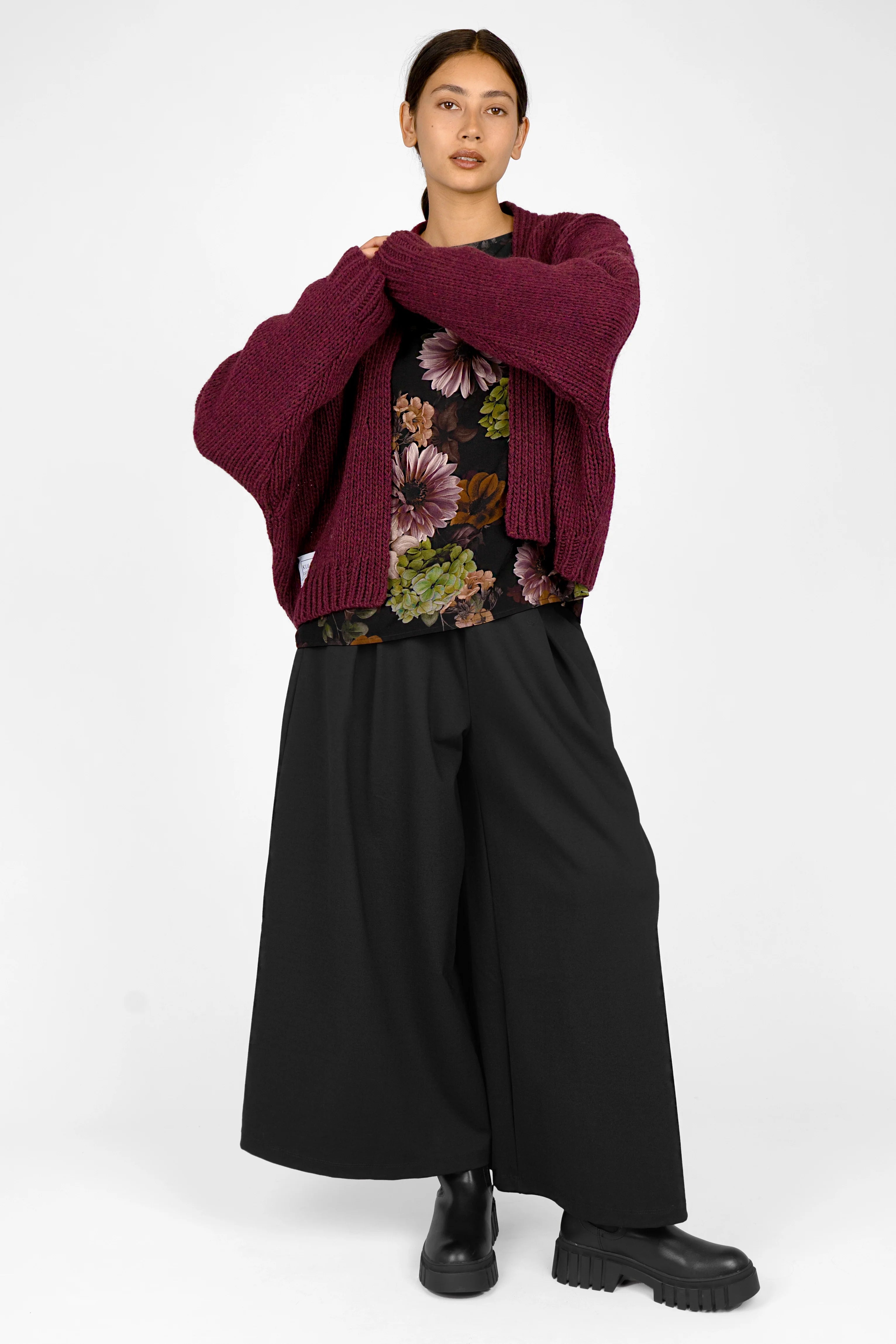 Woman wearing GRETA.K Bluse with allover floral print, paired with burgundy cardigan and black wide-leg pants.